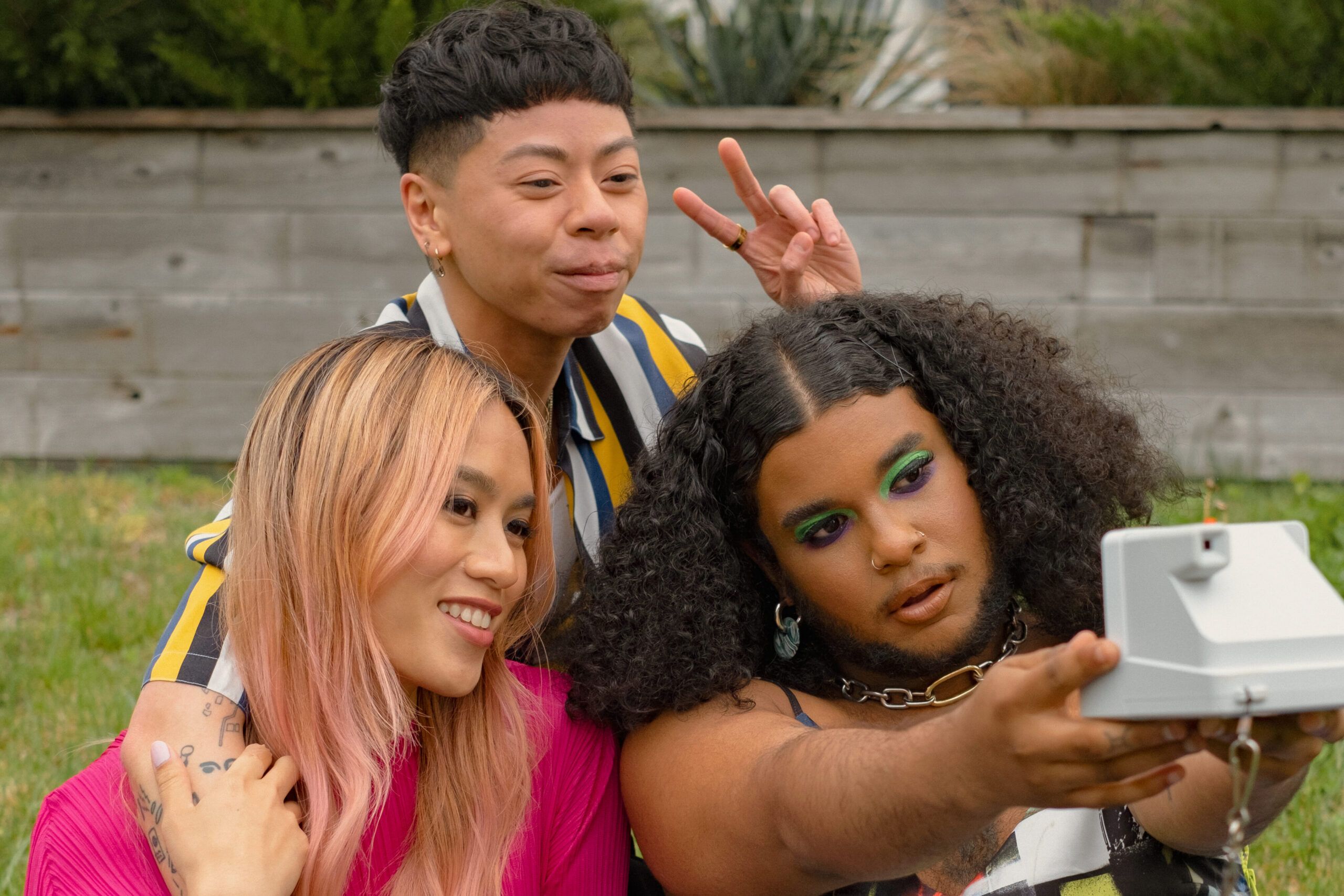 Three friends of varying genders taking a selfie in a park with a polaroid camera