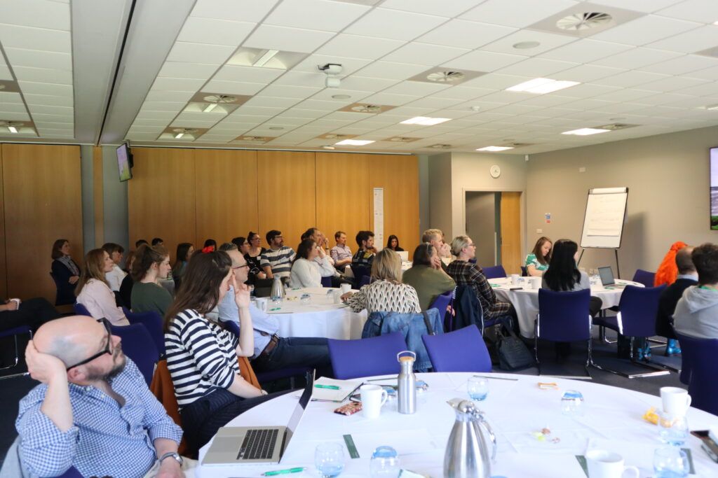 a conference room with groups of people sitting round tables listening to a presentation