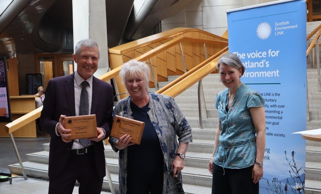 Three people standing next to each other and smiling, two of them holding wooden awards