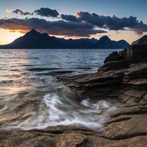 View of a rocky shore at sunset with the waves of the sea swirling over the rocks and mountains across the water.